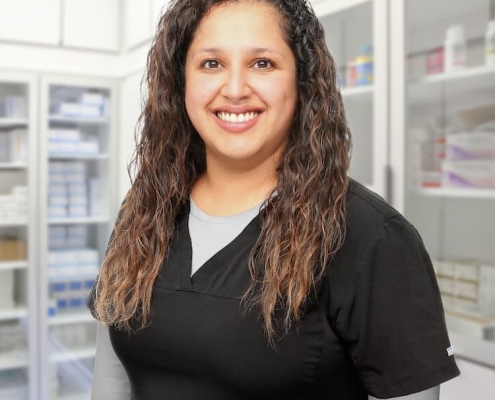 Smiling person with long, wavy hair is wearing a black medical uniform. They are standing in a bright room that appears to be a medical or pharmacy setting, with shelves of various supplies in the background.