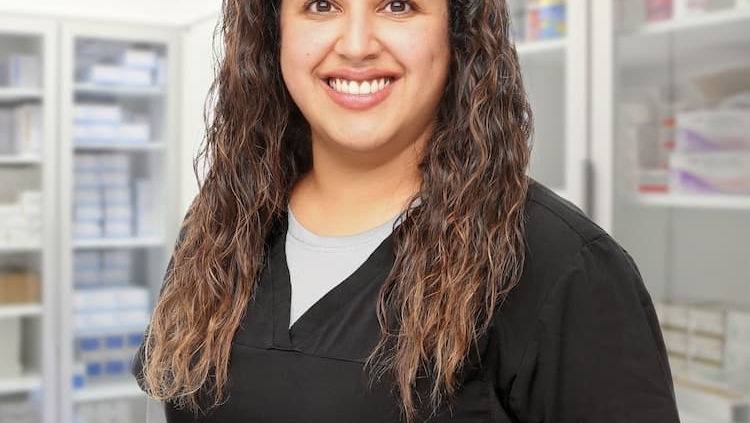 Smiling person with long, wavy hair is wearing a black medical uniform. They are standing in a bright room that appears to be a medical or pharmacy setting, with shelves of various supplies in the background.