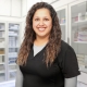 Smiling person with long, wavy hair is wearing a black medical uniform. They are standing in a bright room that appears to be a medical or pharmacy setting, with shelves of various supplies in the background.