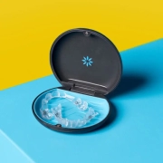 A clear dental aligner is placed inside an open, round dark case with a blue interior. The case rests on a surface divided into two bright colors, yellow and blue, creating a bold contrast.