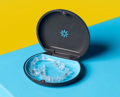 A clear dental aligner is placed inside an open, round dark case with a blue interior. The case rests on a surface divided into two bright colors, yellow and blue, creating a bold contrast.