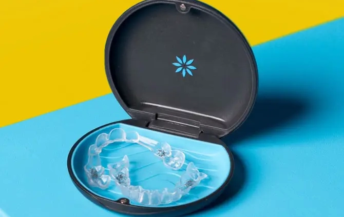 A clear dental aligner is placed inside an open, round dark case with a blue interior. The case rests on a surface divided into two bright colors, yellow and blue, creating a bold contrast.