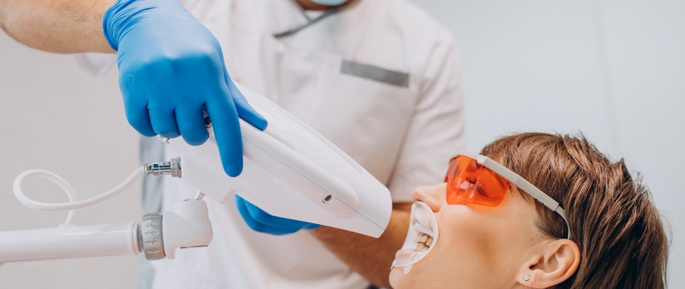 A dentist wearing gloves and a mask uses a whitening device on a patient with protective glasses and a mouthguard in a dental clinic.